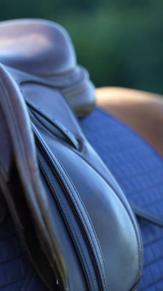 Close up of a brown Cavaletti Collection saddle on a blue quilted pad in an outdoor setting.