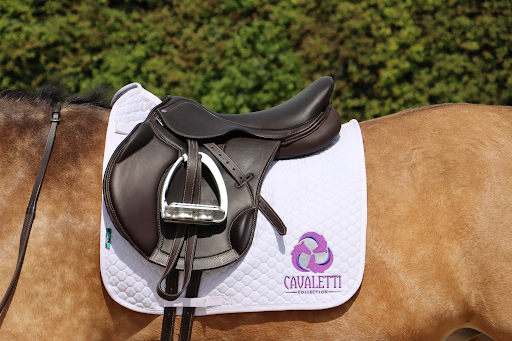 Close‑up of a brown Cavaletti Collection saddle on a white quilted saddle pad in an outdoor setting.