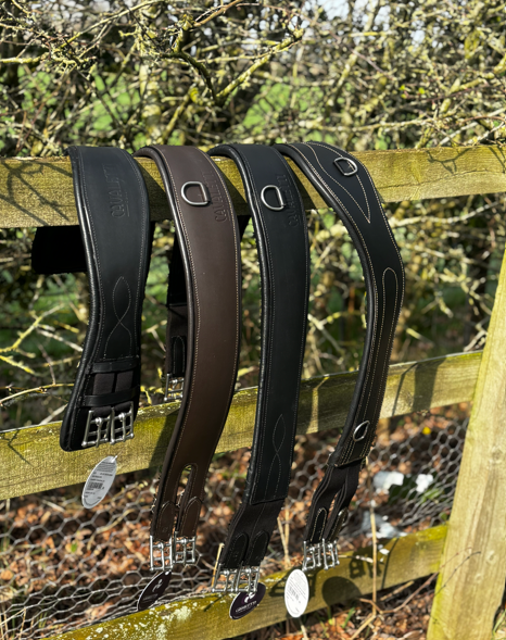 Cavaletti Collection black and brown leather girths hanging over a wooden fence in an outdoor yard setting.