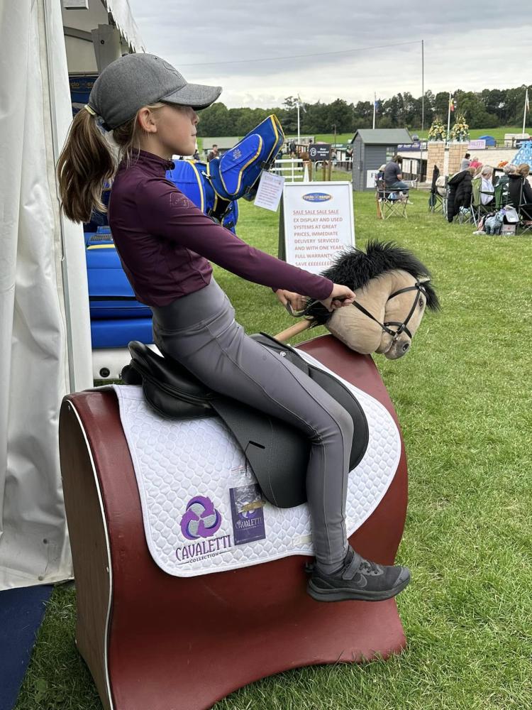 Rider sitting on a mechanical training setup with a white Cavaletti Collection saddle pad during an outdoor event.