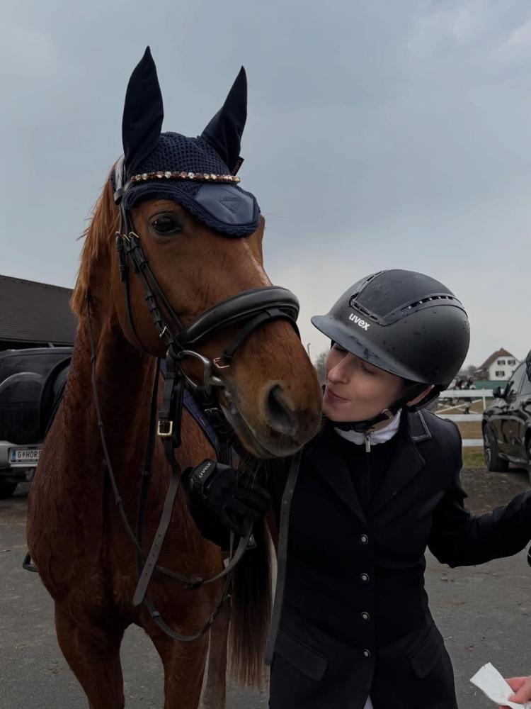 Rider standing beside a brown horse wearing a navy ear bonnet and bridle in an outdoor equestrian setting. Photo credit: vcresch.