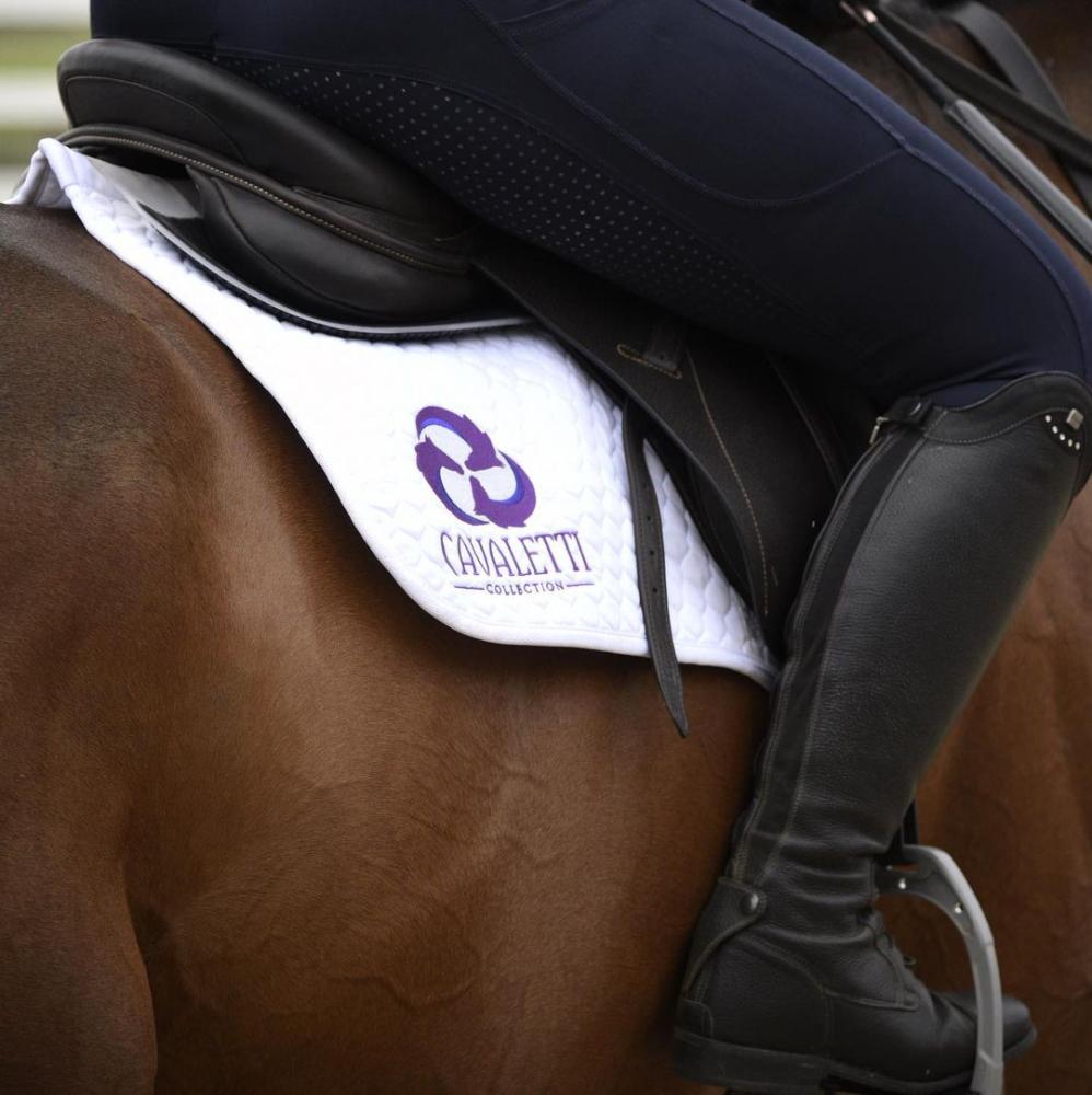 Close up of a riders leg and boot in the stirrup beside a white Cavaletti Collection saddle pad.