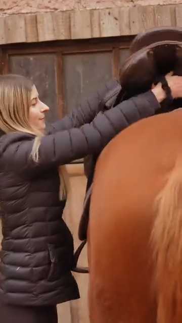 Rider lifting and positioning a saddle onto a horse in a stable yard before riding Cavaletti Collection.