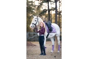 Rider standing beside a white horse fitted with a Cavaletti Monoflap Dressage Saddle and purple saddle pad, showcasing stylish equestrian gear in an outdoor setting.