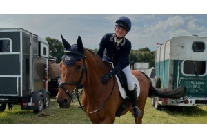Smiling rider in navy show jacket and helmet sitting on a bay horse in tack at an outdoor competition warm up area with horse trailers in the background. Image credit to team_allerton
