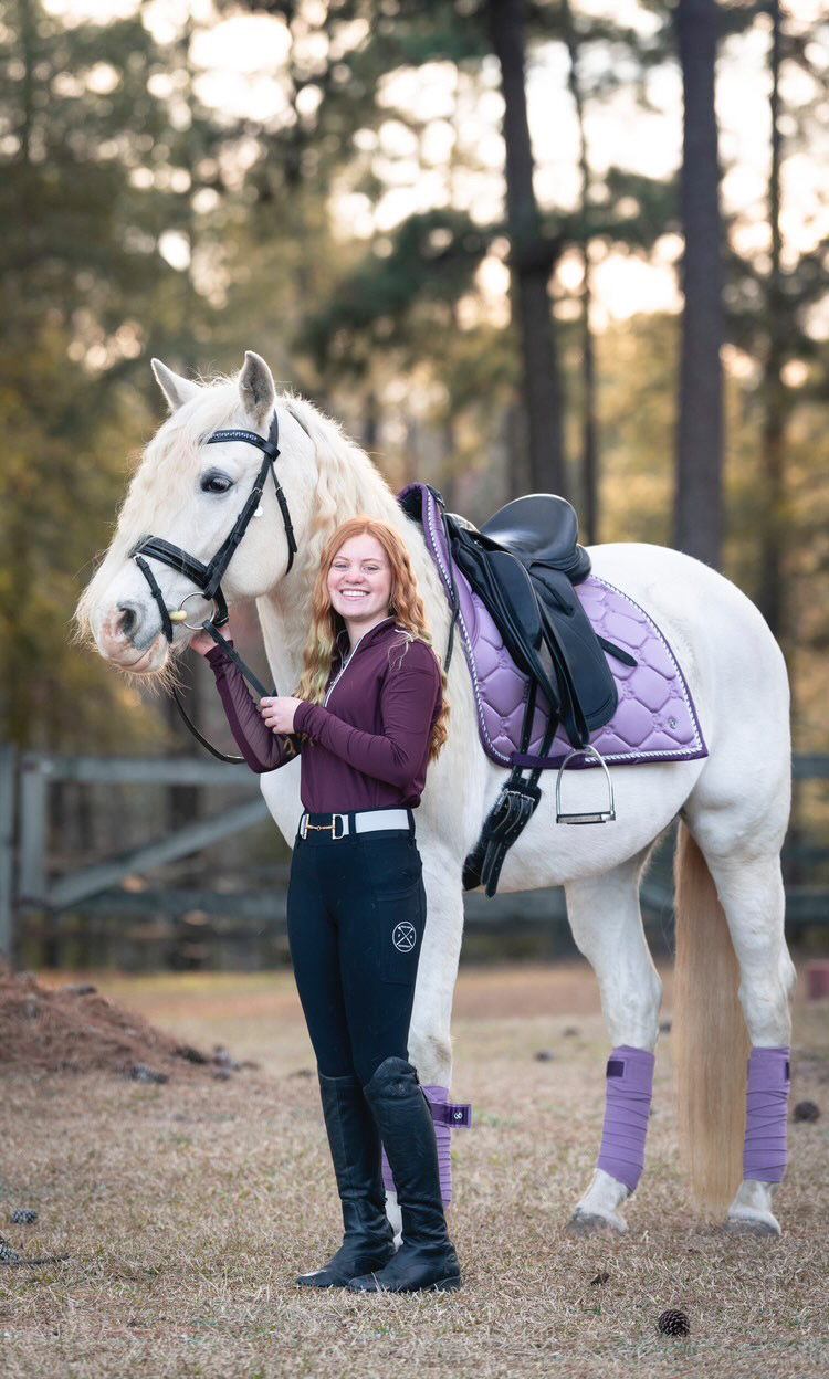 Rider standing beside a white horse fitted with a Cavaletti Monoflap Dressage Saddle and purple saddle pad, showcasing stylish equestrian gear in an outdoor setting.