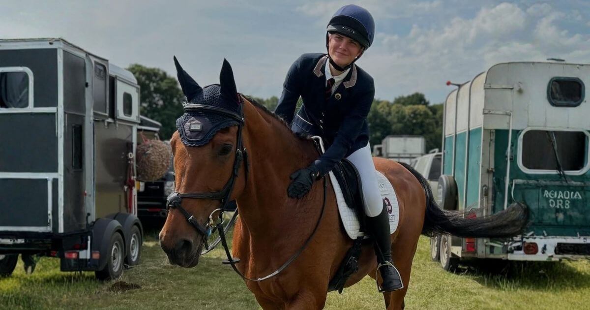 Smiling rider in navy show jacket and helmet sitting on a bay horse in tack at an outdoor competition warm up area with horse trailers in the background. Image credit to team_allerton