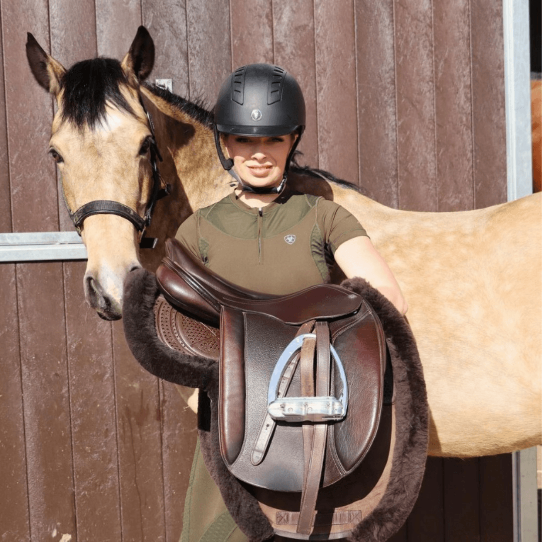 Rider holding a brown leather saddle with sheepskin lining beside a dun horse at the stable door